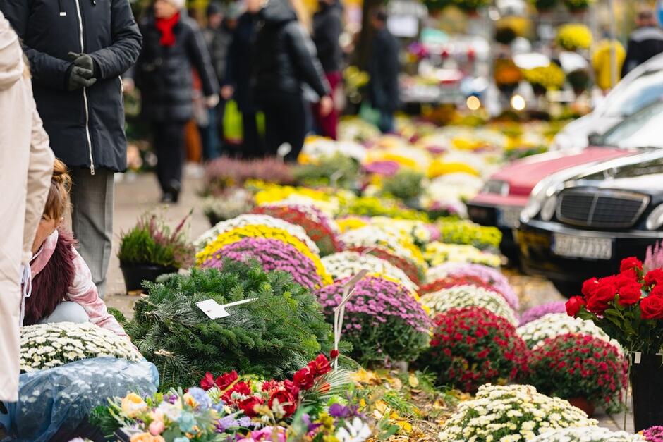 Zdjęcie przedstawia uliczny targ kwiatowy, gdzie na ziemi rozłożone są kolorowe wiązanki kwiatów, głównie chryzantemy, oraz wieńce. W tle widać przechodniów ubranych w zimowe kurtki, a także zaparkowane samochody