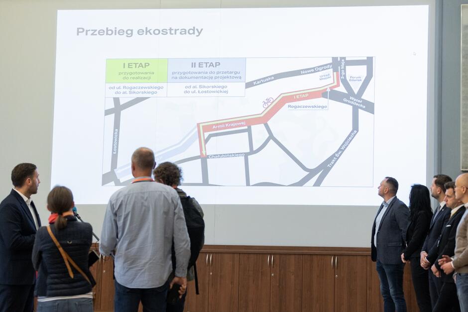 na zdjęciu widać trzech dziennikarzy stoją tyłem do fotografującego nagrywają na mikrofony stojącego przed nimi wiceprezydenta Gdańska, na ścianie wyświetlana jest duża prezentacja