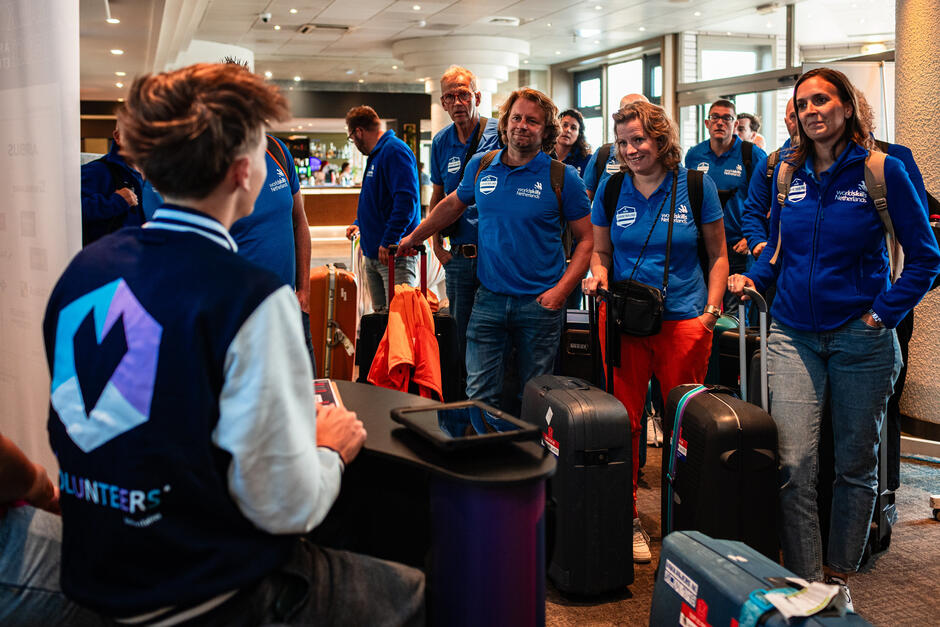 Na zdjęciu widać grupę ludzi ubranych w niebieskie koszulki i kurtki z logo WorldSkills Netherlands,  którzy stoją w kolejce z walizkami, prawdopodobnie podczas rejestracji na wydarzenie lub przy zameldowaniu. Osoba siedząca przy biurku z napisem  Volunteers  obsługuje ich, co sugeruje, że jest to moment akredytacji lub przyjazdu na międzynarodowe wydarzenie.