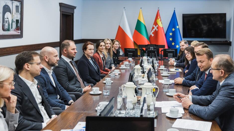 Na zdjęciu widzimy grupę osób siedzących przy długim stole w sali konferencyjnej. Po lewej stronie stołu siedzi siedem osób, a po prawej osiem. Na stole znajdują się filiżanki, dzbanki z wodą i dokumenty. W tle widoczne są flagi Polski, Litwy, Unii Europejskiej oraz czerwono-biała flaga z symbolem miecza i krzyża, która może należeć do jednego z regionów lub miast. Uczestnicy spotkania wyglądają na skoncentrowanych, prawdopodobnie odbywa się dyskusja na temat współpracy międzynarodowej. W pomieszczeniu widać również monitor na ścianie i obraz nawiązujący do architektury.