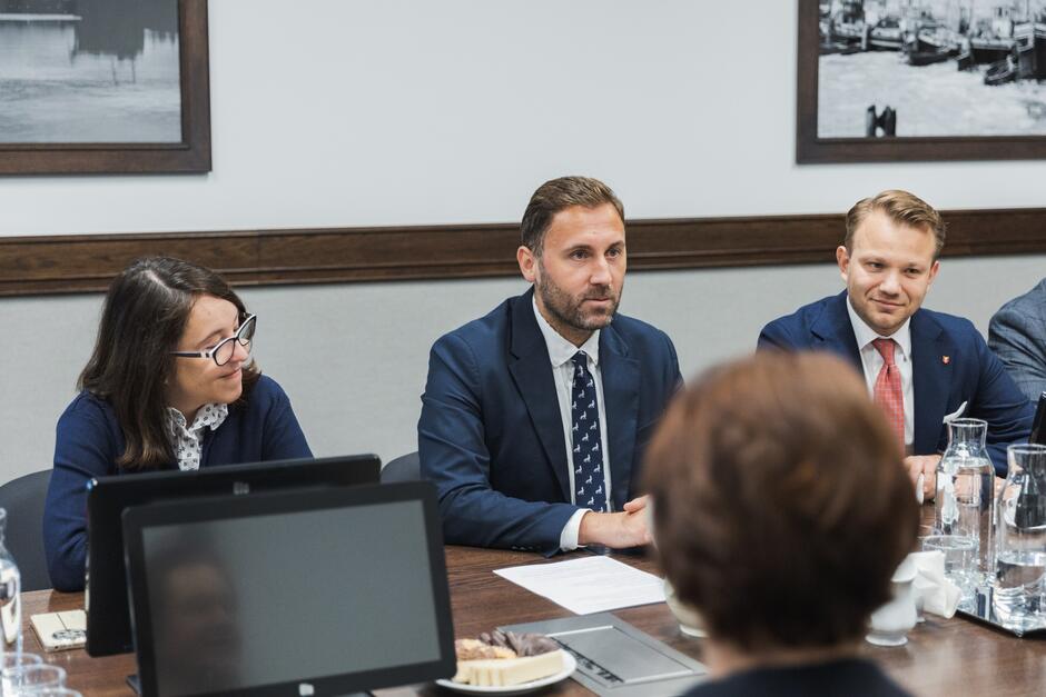 Na zdjęciu widzimy troje uczestników spotkania siedzących przy stole konferencyjnym. Po lewej stronie znajduje się kobieta w okularach, uśmiechnięta, w ciemnym swetrze i koszuli. W centrum siedzi mężczyzna z brodą w eleganckim garniturze, białej koszuli i krawacie w drobne wzory, patrząc uważnie przed siebie. Po jego prawej stronie siedzi inny mężczyzna w ciemnym garniturze, białej koszuli i czerwonym krawacie, który również uśmiecha się delikatnie. Na stole przed nimi znajdują się dokumenty, dzbanki z wodą, filiżanki oraz talerz z ciastkami. W tle widać dwa czarno-białe zdjęcia przedstawiające portowe sceny.