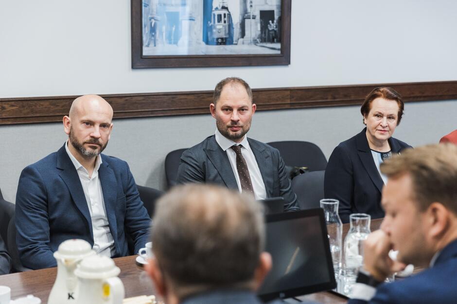 Na zdjęciu widzimy trzy osoby siedzące przy stole konferencyjnym. Po lewej stronie znajduje się mężczyzna z brodą i ogoloną głową, ubrany w ciemny garnitur i białą koszulę. Obok niego siedzi drugi mężczyzna, który ma krótkie włosy i nosi ciemny garnitur, białą koszulę i brązowy krawat w kropki. Po jego prawej stronie siedzi starsza kobieta w ciemnym stroju, patrząca w stronę obiektywu. Na stole przed nimi znajdują się dzbanki, filiżanki oraz szklanki z wodą. W tle wisi obraz przedstawiający uliczną scenę z tramwajem.