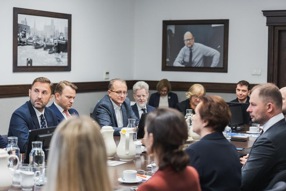 Na zdjęciu widzimy uczestników spotkania siedzących wokół dużego stołu konferencyjnego. Po lewej stronie siedzą trzy osoby: mężczyzna z brodą w granatowym garniturze i krawacie, mężczyzna w ciemnym garniturze z czerwonym krawatem oraz mężczyzna w okularach, ubrany w szary garnitur. Dalej, z tyłu, widać jeszcze kilka osób, w tym dwie starsze kobiety oraz mężczyznę z siwą brodą. Po prawej stronie stołu siedzą inne osoby, ale są odwrócone tyłem lub bokiem. Na stole znajdują się dzbanki, filiżanki, szklanki z wodą i dokumenty. W tle na ścianie widoczne są dwa zdjęcia – jedno przedstawiające portowe nabrzeże, a drugie mężczyznę w okularach i koszuli z podwiniętymi rękawami. Atmosfera spotkania wydaje się formalna.