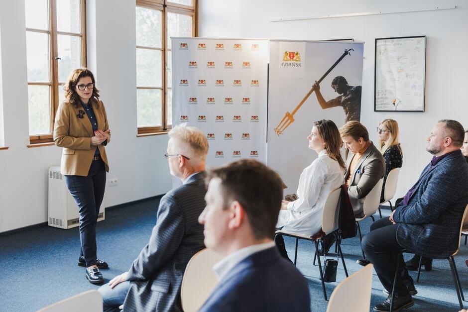 Na zdjęciu widzimy kobietę w marynarce, która przemawia do grupy siedzących osób podczas oficjalnego spotkania, prawdopodobnie związanego z radą zdrowia publicznego w Gdańsku. W tle znajdują się banery z logo Gdańska oraz wizerunkiem Neptuna, co sugeruje, że wydarzenie ma formalny charakter i dotyczy spraw miejskich.