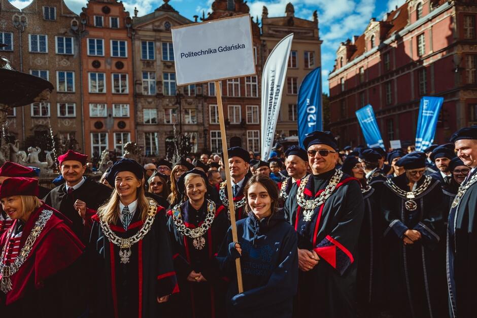 Na zdjęciu widać grupę ludzi uczestniczących w paradzie akademickiej, z przodu znajduje się młoda osoba trzymająca tabliczkę z napisem „Politechnika Gdańska”. W tle widać innych uczestników ubranych w tradycyjne togi akademickie, a sceneria sugeruje, że wydarzenie odbywa się na tle historycznych budynków w Gdańsku