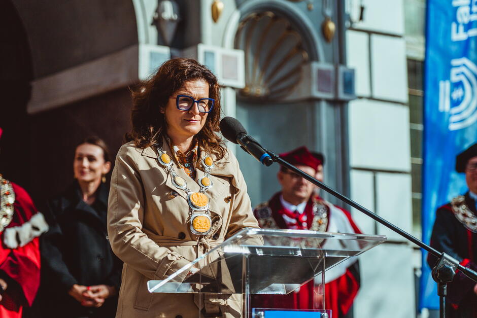 Na zdjęciu widzimy kobietę w okularach, ubranej w płaszcz, przemawiającą przy mównicy na otwartej przestrzeni. W tle stoją osoby w strojach akademickich, co sugeruje, że jest to wydarzenie o charakterze uroczystym, prawdopodobnie inauguracja roku akademickiego