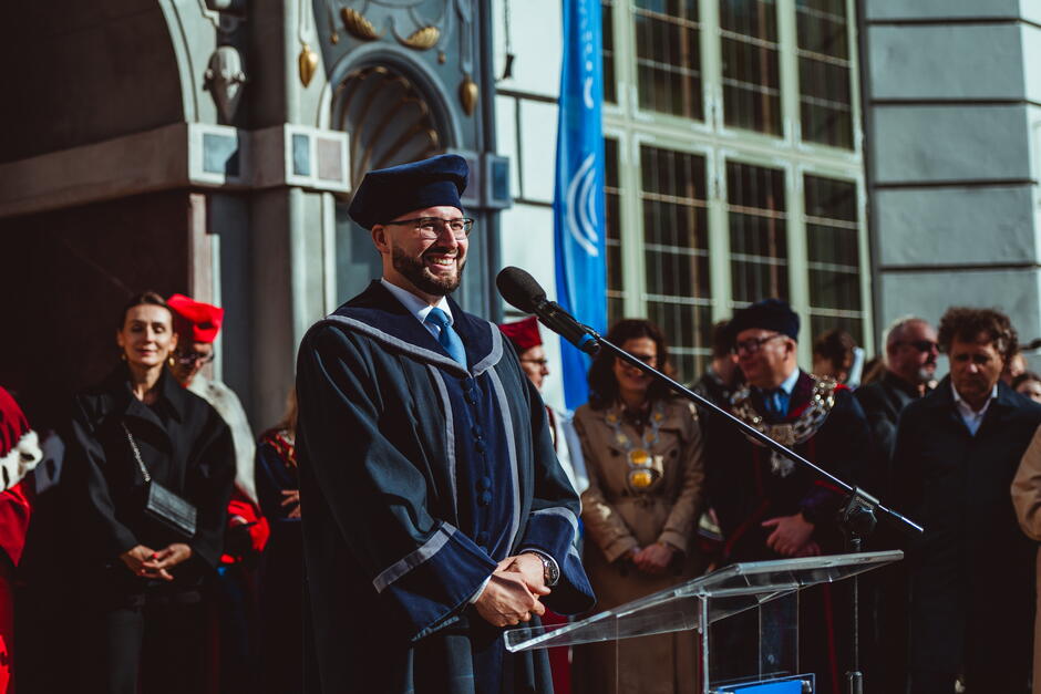 Na zdjęciu widać mężczyznę w stroju akademickim, stojącego przy mównicy i uśmiechającego się podczas przemowy. W tle znajdują się inne osoby, również w togach akademickich, co wskazuje na uroczystość związaną z inauguracją roku akademickiego lub innym oficjalnym wydarzeniem akademickim