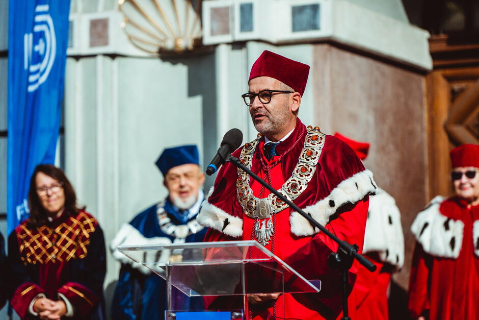 Na zdjęciu widać mężczyznę w czerwonym stroju akademickim i birecie, przemawiającego przy mównicy. Mężczyzna nosi ozdobny łańcuch rektorski, a za nim stoją inne osoby w strojach akademickich, co sugeruje oficjalną uroczystość, prawdopodobnie inaugurację roku akademickiego