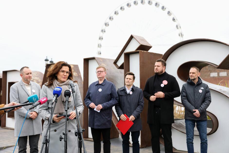 na zdjęciu konferencja prasowa, po lewej stoi kobieta w szarym płaszczu, mówi do mikrofonów dziennikarskich, za nią w jednym rzędzie stoi kilku mężczyzn