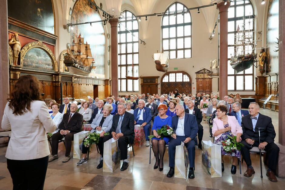 W okazałej, reprezentacyjnej sali siedzi na krzesłach kilkadziesiąt starszych osób, którzy świętują jubileusze małżeńskie. Odwrócona tyłem do aparatu kobieta przemawia do nich. 