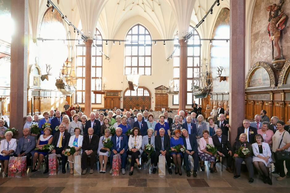 Kilkadziesiąt seniorów pozuje do wspólnej fotografii w okazałym, reprezentacyjnym wnętrzu Dworu Artusa w Gdańsku