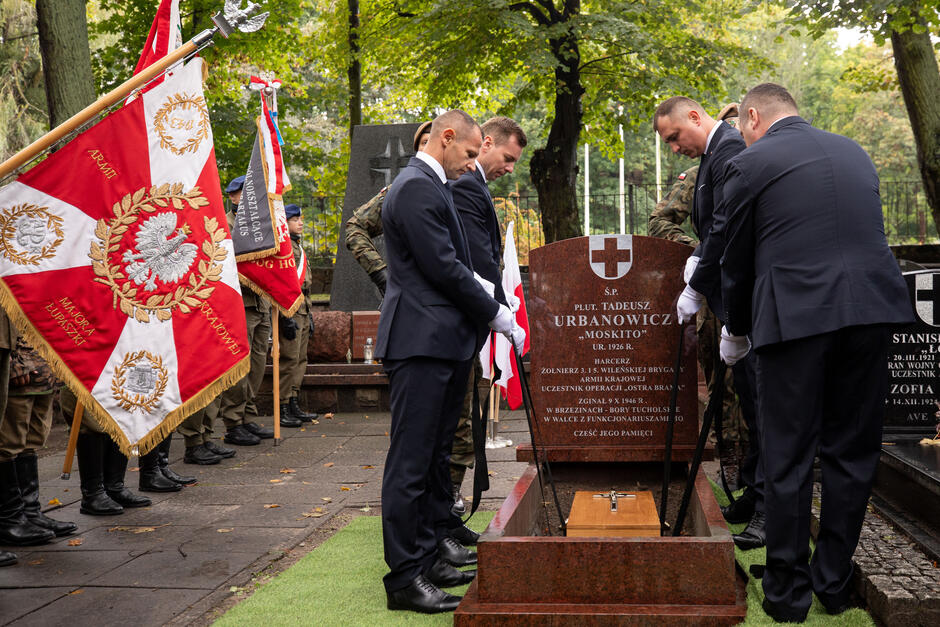 Czterech mężczyzn opuszcza trumnę ze szczątkami do grobu. Po lewej stoi mężczyzna z biało-czerwony sztandarem typu wojskowego 