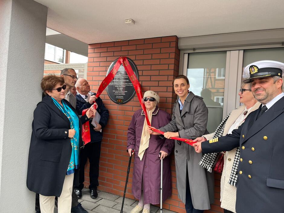 moment uroczystego odsłonięcia tablica, przy czarnej tablicy na ścianie stoi kilka dorosłych osób, dwie z nich trzymają w ręce czerwoną wstęgę