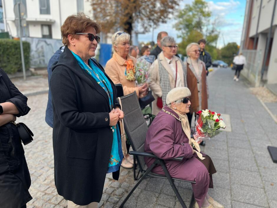 na zdjęciu kilka starszych osób, w ciepłej jesiennej odzieży, większość stoi i patrzy przed siebie, jedna starsza pani siedzi na krześle