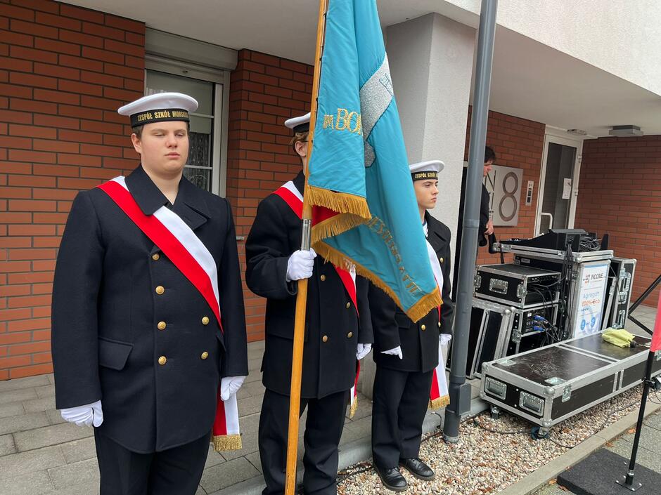 na zdjęciu trzech uczniów szkoły średniej, w marynarskich strojach, ten po środku trzyma sztandar