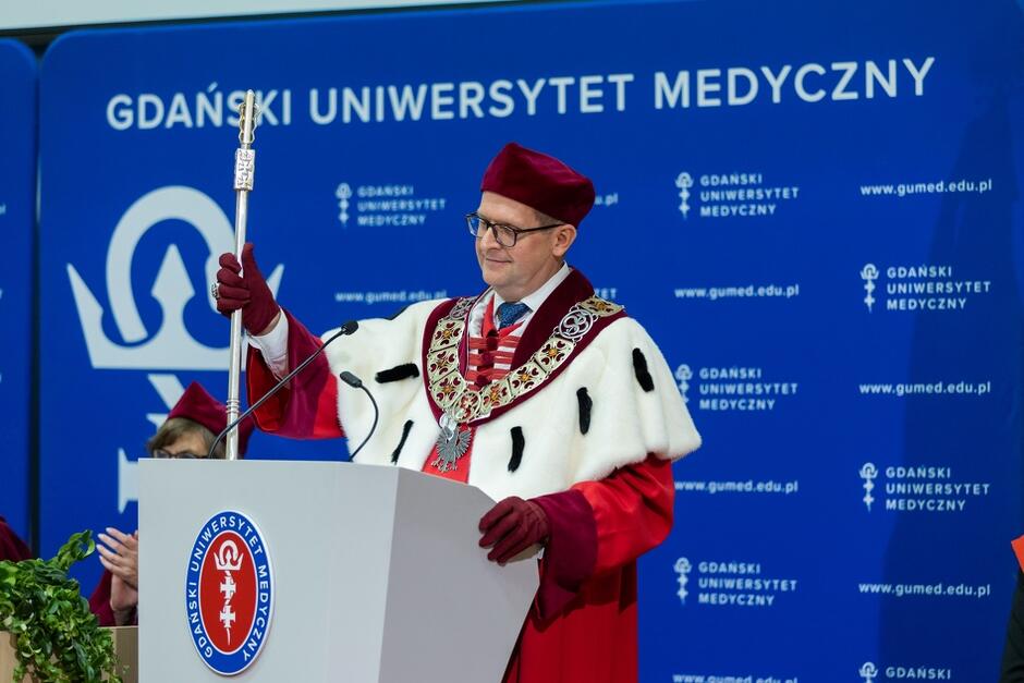 Na zdjęciu widzimy mężczyznę w ceremonialnej todze i birecie, stojącego przy mównicy z insygniami w ręku, podczas oficjalnej uroczystości na Gdańskim Uniwersytecie Medycznym. Jego strój oraz berło sugerują, że jest to rektor lub inna wysoka osoba funkcyjna, a wydarzenie ma charakter akademicki, prawdopodobnie inauguracja roku akademickiego.