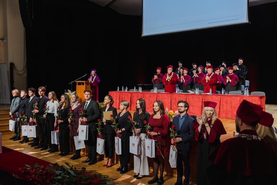 Na zdjęciu widzimy grupę młodych osób stojących na scenie, trzymających kwiaty oraz torby z upominkami. W tle, za stołem, zasiadają osoby w togach akademickich, a w głębi jedna z nich przemawia z mównicy