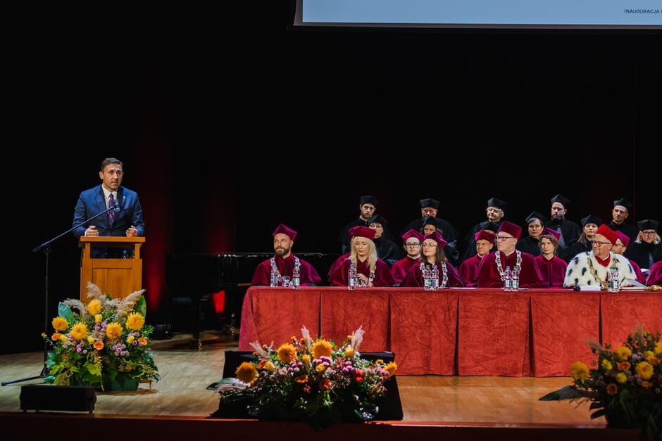 Na zdjęciu widać mężczyznę w garniturze przemawiającego z mównicy podczas uroczystości akademickiej, a na scenie przed nim znajdują się osoby w togach akademickich, zasiadające za stołem przykrytym czerwonym obrusem. W tle można zauważyć więcej osób ubranych w czarne togi, a przed sceną ustawione są ozdobne kompozycje kwiatowe.