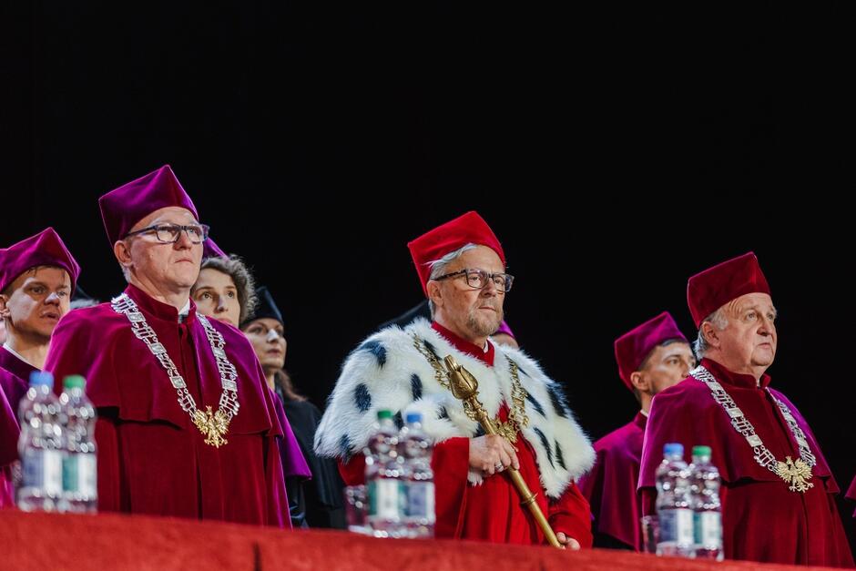 Na zdjęciu widzimy grupę osób w togach i biretach akademickich, stojących za stołem podczas uroczystości, z przodu trzech mężczyzn w czerwonych togach. Jeden z nich, ubrany w togę obszytą futrem, trzyma ceremonialną laskę - pełni funkcję rektora