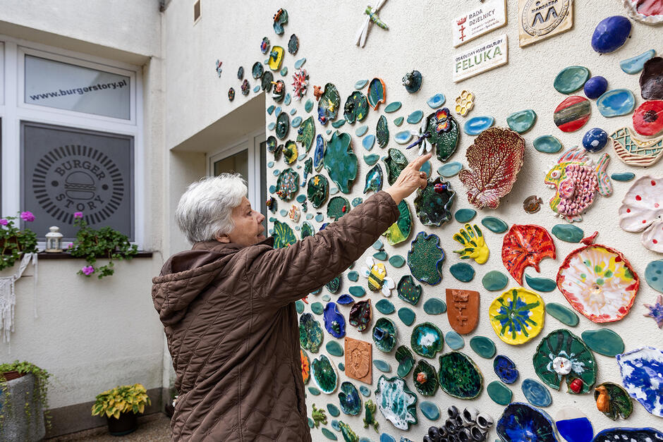 Starsza kobieta stoi przed ścianą wyłożona pracami ceramicznymi i wskazuje na jedną z nich.