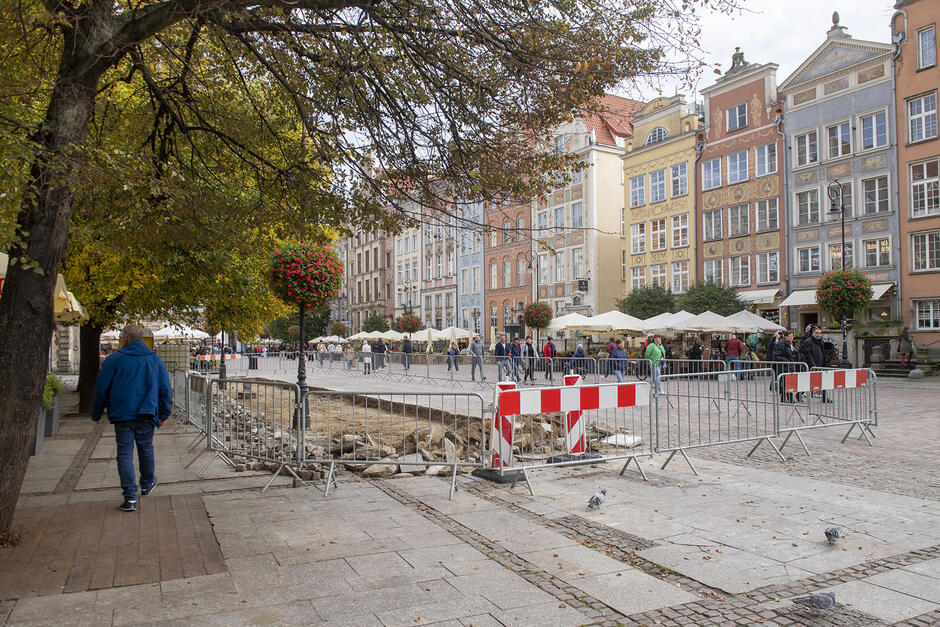 Na zdjęciu widzimy część ulicy w Gdańsku, gdzie trwają prace remontowe, a teren jest odgrodzony barierkami. W tle znajdują się zabytkowe kamienice i spacerujący ludzie, a także ogródki restauracyjne pod parasolami, co sugeruje, że prace odbywają się w popularnej części miasta.