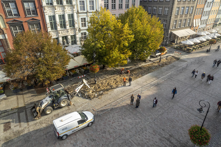 Na zdjęciu widzimy fragment ulicy w centrum miasta, prawdopodobnie Gdańska, gdzie trwają prace drogowe, w tym wykop oraz sprzęt budowlany, taki jak koparka. Obok remontowanej części ulicy spacerują ludzie, a w tle widać kamienice oraz ogródki restauracyjne z parasolami.