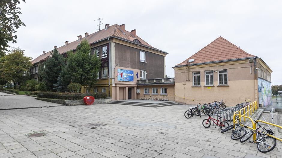 Na zdjęciu widoczny jest budynek szkolny z rowerami zaparkowanymi przed nim. Budynek ma kilka pięter i brązowy dach. Na fasadzie budynku znajduje się napis „Przedszkole nr 17” oraz duży baner reklamowy. Przed szkołą widać stojaki na rowery, a przy nich zaparkowane są różne rowery. Na placu jest duża przestrzeń wyłożona kostką brukową. W tle rosną drzewa, które dodają zieleni otoczeniu. Budynek po prawej stronie ma mural na jednej ze ścian. Cała scena sprawia wrażenie spokojnego otoczenia szkolnego, być może w godzinach popołudniowych.