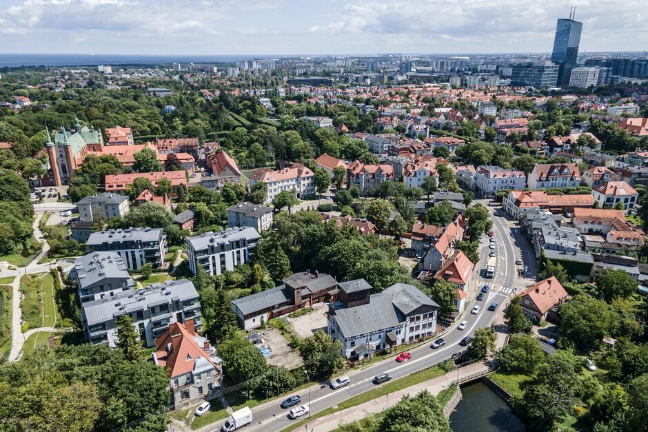 zdjęcie z drona, widać niewysokie kamienice, częściowo z czerwonymi dachami, w tle po lewej kościół po prawej w tle wysoki szklany biurowiec, między budynkami dużo zielonych drzew