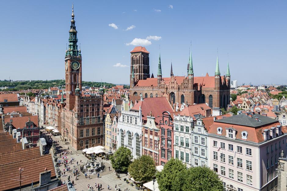 Na zdjęciu widzimy plac w centrum Gdańska z widokiem na Ratusz Głównego Miasta oraz Fontannę Neptuna, otoczony zabytkowymi kamienicami i budynkiem Dworu Artusa. Na placu znajduje się wielu turystów i spacerowiczów, a pod parasolami widoczne są kawiarnie, co wskazuje na popularność tego miejsca wśród odwiedzających.