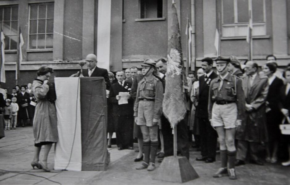 Zdjęcie przedstawia oficjalną uroczystość, prawdopodobnie związaną z harcerstwem, odbywającą się na zewnątrz, na placu przed budynkiem. Na pierwszym planie widzimy mężczyznę przemawiającego przy mównicy ozdobionej biało-czerwoną flagą. Obok niego stoi kobieta, która wydaje się brać udział w ceremonii. Po prawej stronie zdjęcia stoją harcerze w pełnym umundurowaniu, trzymający sztandar z wizerunkiem orła, co podkreśla patriotyczny charakter wydarzenia. Za nimi widać grupę dorosłych, którzy przyglądają się ceremonii. Niektórzy z nich mają przypięte odznaki, co sugeruje, że mogą pełnić oficjalne role. W tle widoczny jest tłum zgromadzonych osób, w tym dzieci oraz kilku mężczyzn w garniturach, co wskazuje na uroczysty charakter tego wydarzenia. Na budynku widać również powiewające flagi, co dodatkowo podkreśla oficjalność spotkania.  Zdjęcie jest czarno-białe i pochodzi prawdopodobnie z okresu PRL-u. Atmosfera na zdjęciu jest formalna, a cała scena wskazuje na znaczącą uroczystość harcerską lub szkolną.