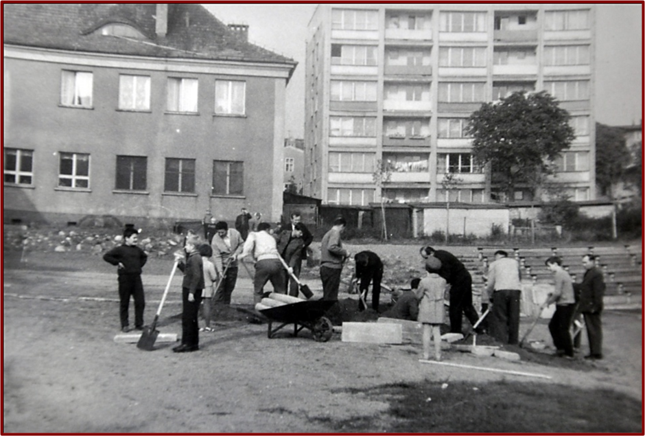 Zdjęcie przedstawia grupę ludzi pracujących na świeżym powietrzu, prawdopodobnie przy budowie lub renowacji placu lub boiska szkolnego. Na pierwszym planie widzimy mężczyzn, młodzież i dzieci zaangażowanych w prace fizyczne. Część z nich trzyma łopaty, inni przenoszą materiały, jak cegły czy betonowe elementy. Na środku widoczna jest taczka, której używają do transportu materiałów budowlanych. Prace odbywają się na placu przed budynkiem, który wygląda na szkołę lub inny publiczny budynek, o prostych kształtach i dużych oknach. W tle, po prawej stronie, widoczny jest wielopiętrowy blok mieszkalny, typowy dla czasów PRL-u, co sugeruje, że scena rozgrywa się w mieście lub na osiedlu.  Ludzie pracują w zorganizowany sposób, co może wskazywać na czyn społeczny, typowy dla tamtego okresu. Atmosfera jest spokojna, a uczestnicy skupieni na swoich zadaniach. Cała scena utrzymana jest w czarno-białej tonacji, co sugeruje, że zdjęcie pochodzi z połowy XX wieku.