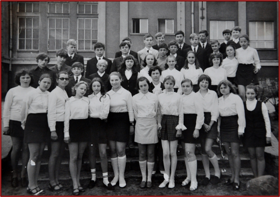 Zdjęcie przedstawia grupę uczniów, prawdopodobnie klasy szkolnej, pozujących do wspólnej fotografii na zewnątrz, przed budynkiem szkoły. Wszyscy uczniowie są elegancko ubrani, co sugeruje, że zdjęcie zostało zrobione podczas oficjalnej okazji, takiej jak zakończenie roku szkolnego lub inna uroczystość. Dziewczęta mają na sobie białe bluzki i spódnice, a niektóre noszą podkolanówki. Chłopcy są ubrani w garnitury lub marynarki z krawatami. Grupa stoi na schodach, a za nimi w tle widoczny jest budynek szkoły o prostych, surowych ścianach z dużymi oknami, typowymi dla architektury szkolnej w okresie PRL.  Na zdjęciu można dostrzec różnorodność fryzur i postaw uczniów, niektóre dziewczęta są przytulone, a inni stoją swobodnie. Atmosfera wydaje się być formalna, ale jednocześnie swobodna, z lekkimi uśmiechami na twarzach uczniów. Fotografia utrzymana jest w czarno-białej kolorystyce, co sugeruje, że została zrobiona w latach 70.