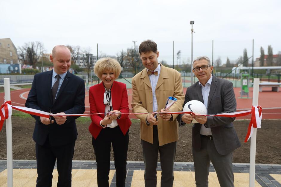 Zdjęcie przedstawia uroczystość otwarcia nowego obiektu sportowego, najprawdopodobniej boiska szkolnego. Czworo elegancko ubranych osób, trzyma nożyczki, przecinając wstęgę, co jest symbolicznym gestem otwarcia. Osoby na zdjęciu są uśmiechnięte, co wskazuje na radosny charakter wydarzenia. Jedna z kobiet ubrana jest w czerwony żakiet, a mężczyzna obok niej trzyma w rękach piłkę, co dodatkowo podkreśla sportowy kontekst uroczystości. W tle widać boisko z bieżnią oraz urządzenia do ćwiczeń na świeżym powietrzu, co sugeruje, że nowo otwarty kompleks jest przeznaczony do różnych aktywności sportowych. Cała scena rozgrywa się na terenie szkoły lub placówki edukacyjnej, a obecność flagi oraz formalnych strojów sugeruje, że w uroczystości biorą udział osoby o ważnych funkcjach, np. dyrekcja szkoły lub przedstawiciele władz lokalnych.