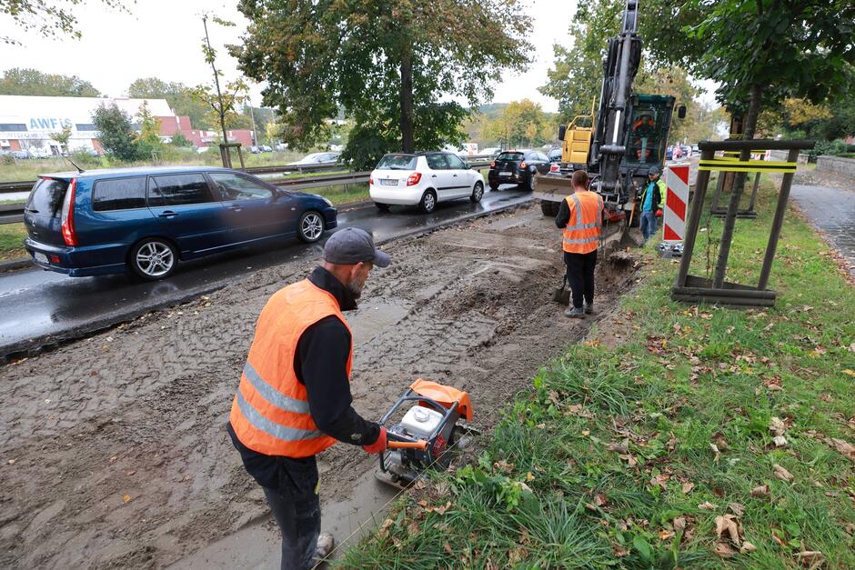 na zdjęciu robotnik w pomarańczowej kamizelce, pcha sprzęt do utwardzania piasku, w tle inny pracownik w kamizelce, jest też rozkopany pas jezdni, a w tle samochody jadące normalną jezdnią