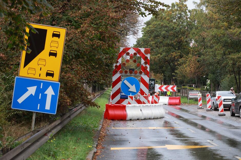 fragment ulicy z asfaltem, widać oznakowanie drogowe, wraz z biało czerwonymi barierami, informujące o zwężeniu jezdni