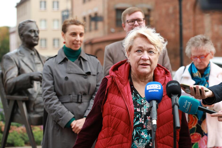 Na zdjęciu znajduje się kobieta w czerwonej kamizelce, która przemawia do mikrofonów z logo stacji TVP3 Gdańsk oraz Radio Gdańsk. Wygląda na zaangażowaną w wypowiedź, a za nią stoją inne osoby: kobieta w szarym płaszczu, mężczyzna w garniturze oraz starsza kobieta w jasnym płaszczu. W tle widać pomnik Lecha Bądkowskiego. Scena odbywa się na zewnątrz, w miejskiej przestrzeni, a ludzie wydają się uczestniczyć w uroczystości lub wydarzeniu medialnym.