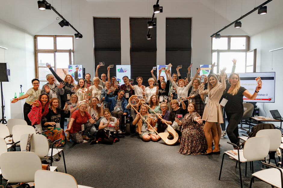 Na zdjęciu znajduje się grupa około 35 osób, które z entuzjazmem pozują do wspólnej fotografii w jasnym pomieszczeniu. Część z nich trzyma kartki lub zdjęcia, a atmosfera wydaje się radosna i pełna pozytywnej energii.