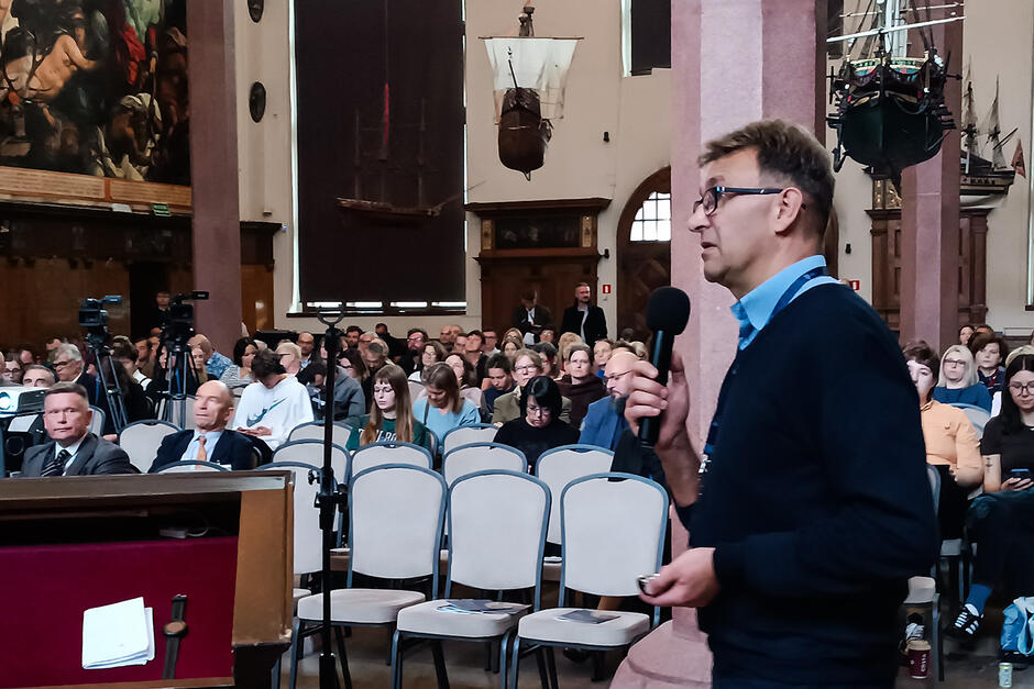 W okazałej sali mężczyzna mówi do mikrofonu, za nim siedzą ludzie na krzesłach. 