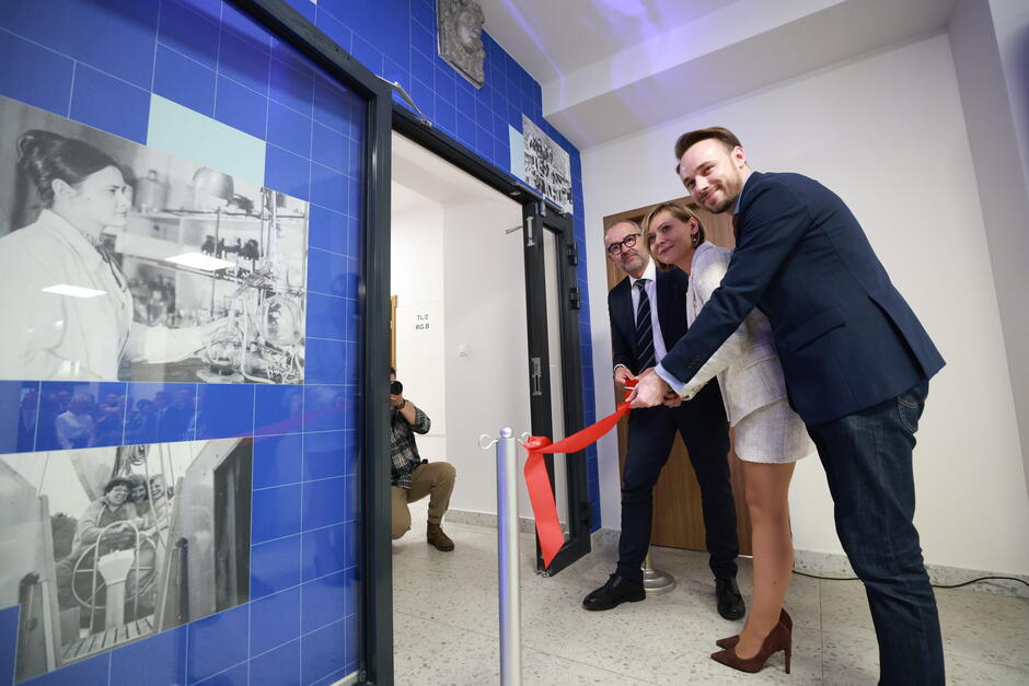 Na zdjęciu widzimy trójkę elegancko ubranych osób przecinających czerwoną wstęgę, symbolicznie otwierającą nowe pomieszczenie. W tle znajdują się czarno-białe fotografie przedstawiające naukowców podczas pracy w laboratorium, a także fotograf z aparatem, dokumentujący to wydarzenie.