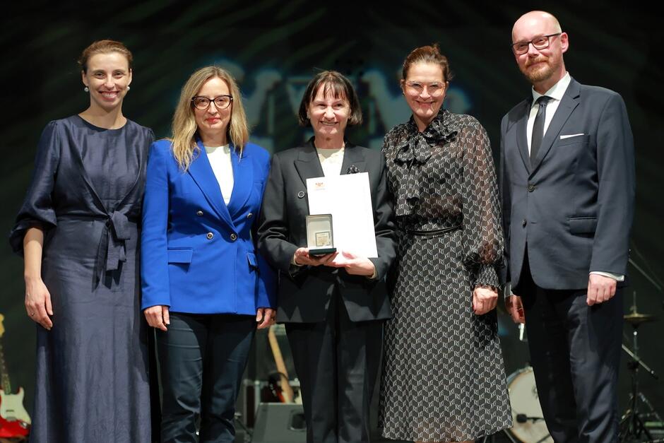 Na zdjęciu widzimy grupę pięciu osób, z których jedna trzyma medal i dyplom, co sugeruje, że została uhonorowana nagrodą. Wszyscy ubrani są formalnie, a zdjęcie pochodzi z uroczystości wręczenia wyróżnień, prawdopodobnie dla nauczycieli.