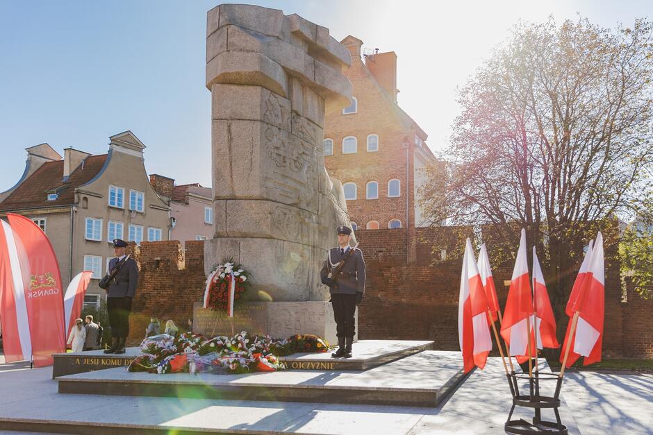 Na zdjęciu widać pomnik w Gdańsku upamiętniający bohaterów poległych za ojczyznę, przy którym stoją dwaj żołnierze w galowych mundurach pełniący wartę honorową. Wokół pomnika widoczne są liczne wieńce i biało-czerwone flagi, co sugeruje uroczystość patriotyczną, prawdopodobnie związaną z obchodami narodowego święta.