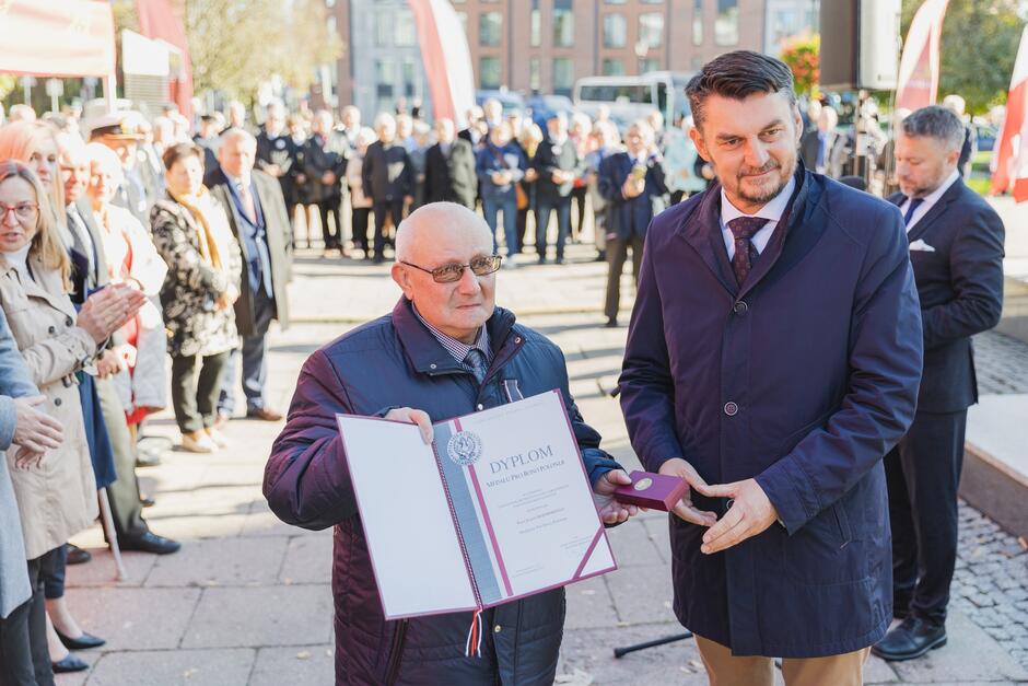 Na zdjęciu dwóch mężczyzn uczestniczy w oficjalnej uroczystości wręczenia odznaczeń. Jeden z nich trzyma dyplom oraz medal, podczas gdy drugi, w ciemnym płaszczu, wręcza mu te odznaczenia; w tle widać grupę osób, która obserwuje to wydarzenie.