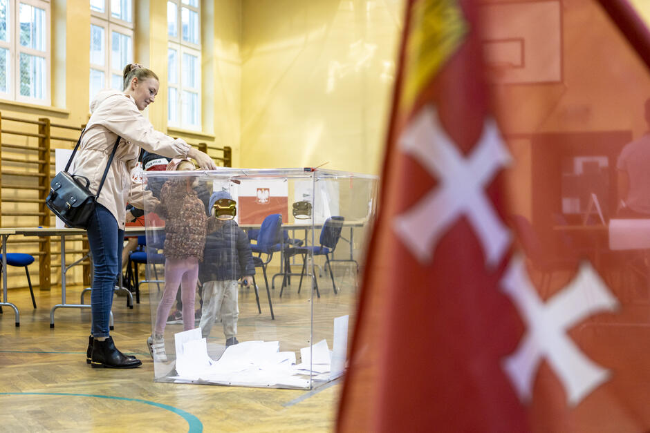 kobieta i dwoje małych dzieci stoją przy urnie i wrzucają do niej kartkę. Cała sytuacja sfotografowana jest we wnętrzu sportowej sali. Na pierwszym planie flaga z dwoma krzyżami i koroną nad nimi