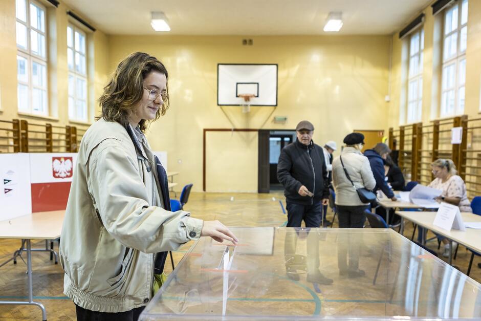 Kobieta wrzuca kartkę do urny wyborczej. W tle inne osoby