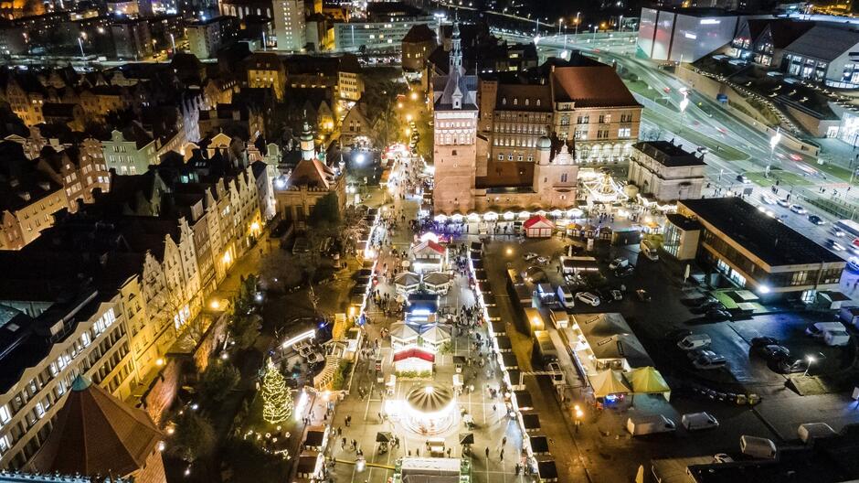 Zdjęcie przedstawia widok z lotu ptaka na oświetlony jarmark bożonarodzeniowy w mieście nocą. Centralną część zajmuje plac z szeregiem stoisk handlowych, rozświetlonych światełkami, oraz karuzelą. Na pierwszym planie widoczna jest udekorowana choinka, a dalej rozpościera się historyczna zabudowa miasta z charakterystycznymi, stromymi dachami i kamienicami. W tle widać większe budynki oraz nowoczesną infrastrukturę drogową. Cała scena tworzy świąteczną atmosferę z dużą ilością świateł i odwiedzających.