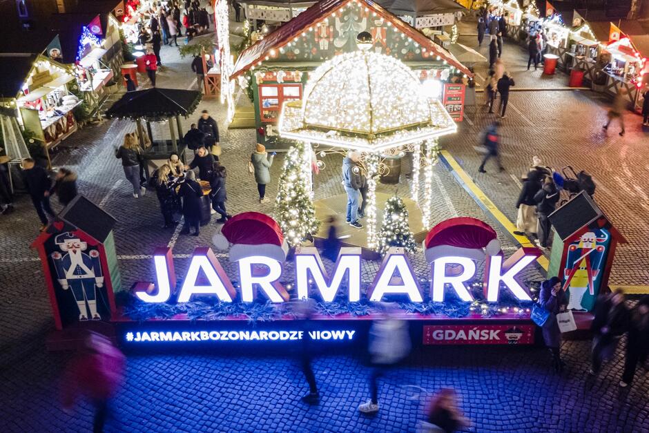 Zdjęcie przedstawia scenę z jarmarku bożonarodzeniowego, prawdopodobnie w Gdańsku, o czym świadczy widoczny napis „GDAŃSK” w prawym dolnym rogu. Na pierwszym planie widać duży, podświetlony napis „JARMARK”, ozdobiony świątecznymi elementami, takimi jak czapki Mikołaja. W tle znajdują się świąteczne stoiska, oświetlone lampkami, które sprzedają różnorodne produkty i jedzenie. Dekoracje wokół są utrzymane w świątecznym klimacie – na zdjęciu można dostrzec małe, oświetlone choinki oraz duży pawilon ozdobiony białymi lampkami. Ludzie spacerują po jarmarku, ciesząc się atmosferą.