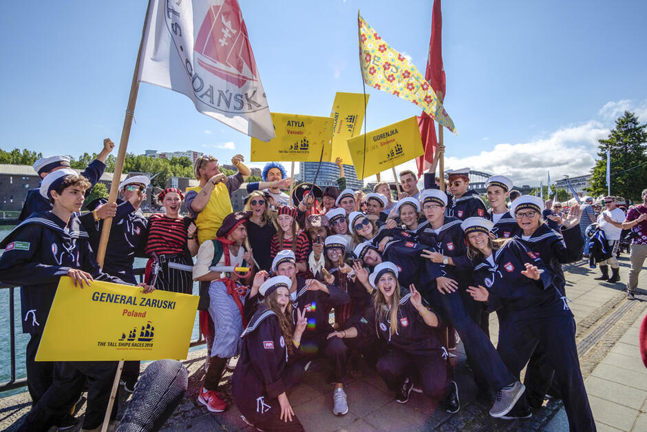 Na zdjęciu widać grupę młodych ludzi ubranych w marynarskie mundury, uczestniczących w paradzie lub święcie związanym z The Tall Ships Races. Trzymają tablice z nazwami statków, takie jak Generał Zaruski  i  Atyla,  oraz flagi, a ich radosne uśmiechy i pozy podkreślają atmosferę entuzjazmu i wspólnoty.