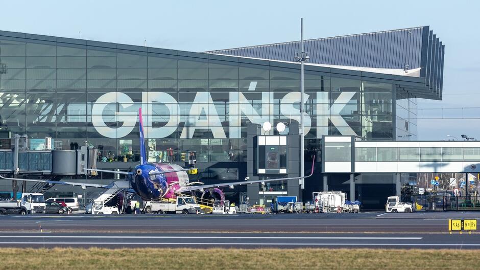 Na zdjęciu znajduje się terminal lotniska w Gdańsku (Gdańsk Airport). W tle widać nowoczesny budynek z dużymi, przeszklonymi ścianami, na których widnieje napis GDAŃSK . Na pierwszym planie znajduje się samolot w barwach fioletowo-niebieskich, który jest obsługiwany przez personel lotniska. Na płycie lotniska widoczne są także różne pojazdy lotniskowe i schody do samolotu. Pogoda wydaje się być słoneczna, a wszystko wskazuje na nowoczesną i dobrze zorganizowaną infrastrukturę lotniska.