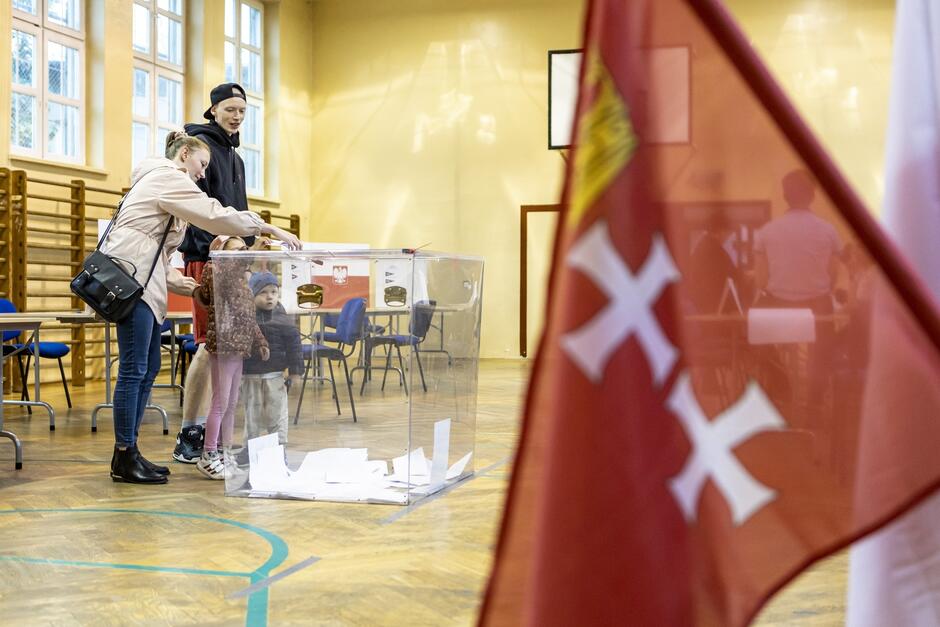 Na zdjęciu widać rodzinę wrzucającą kartę do przezroczystej urny wyborczej w lokalu, który wygląda na salę gimnastyczną. Na pierwszym planie widoczna jest flaga z symbolem krzyża, a w tle znajdują się stoliki i krzesła, co wskazuje, że trwa proces głosowania, prawdopodobnie w Polsce.