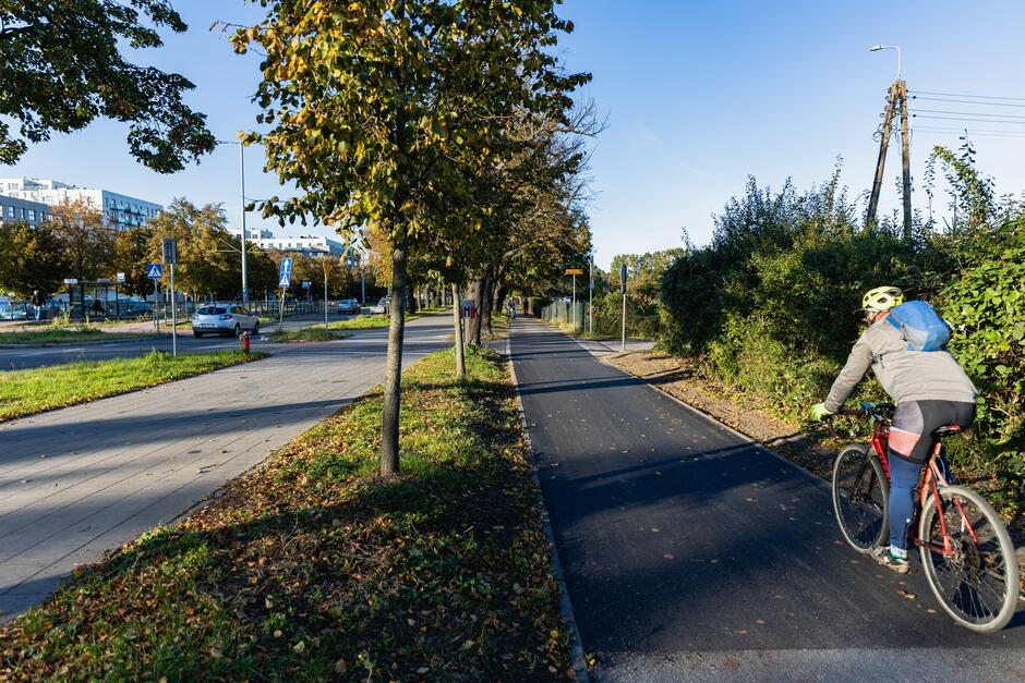 na zdjęciu po prawej wyremontowana droga rowerowa o czarnej nawierzchni, jedzie po niej rowerzysta z kaskiem na głowie, obok drogi rosną w szeregu zielone drzewa, w tle jezdnia samochodowa